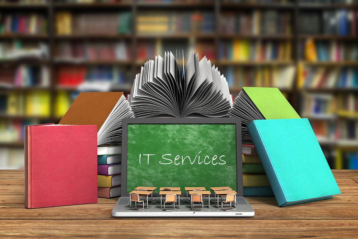 A laptop sits on a desk in a room full of books. The laptop screen is showing a green chalkboard with the words "IT Services" written in chalk.