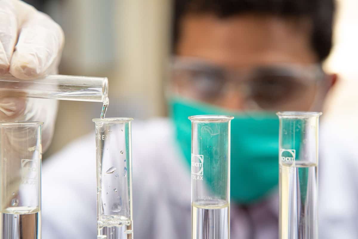 A laboratory technician in gloves and a mask carefully pours liquid from a test tube into another test tube, surrounded by several glass containers filled with clear liquids.