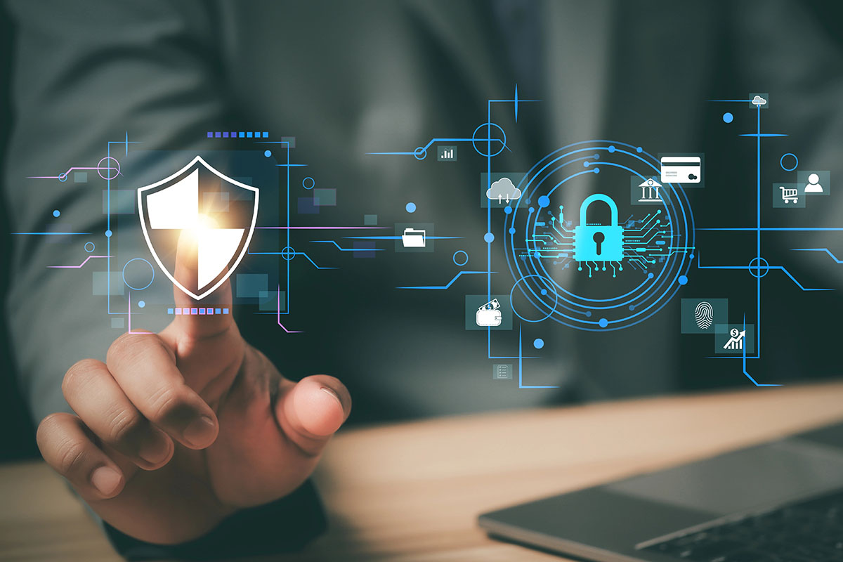A close-up of a hand reaching towards a digital shield symbol, surrounded by cybersecurity graphics, including a padlock and various data icons, with a laptop visible in the background.
