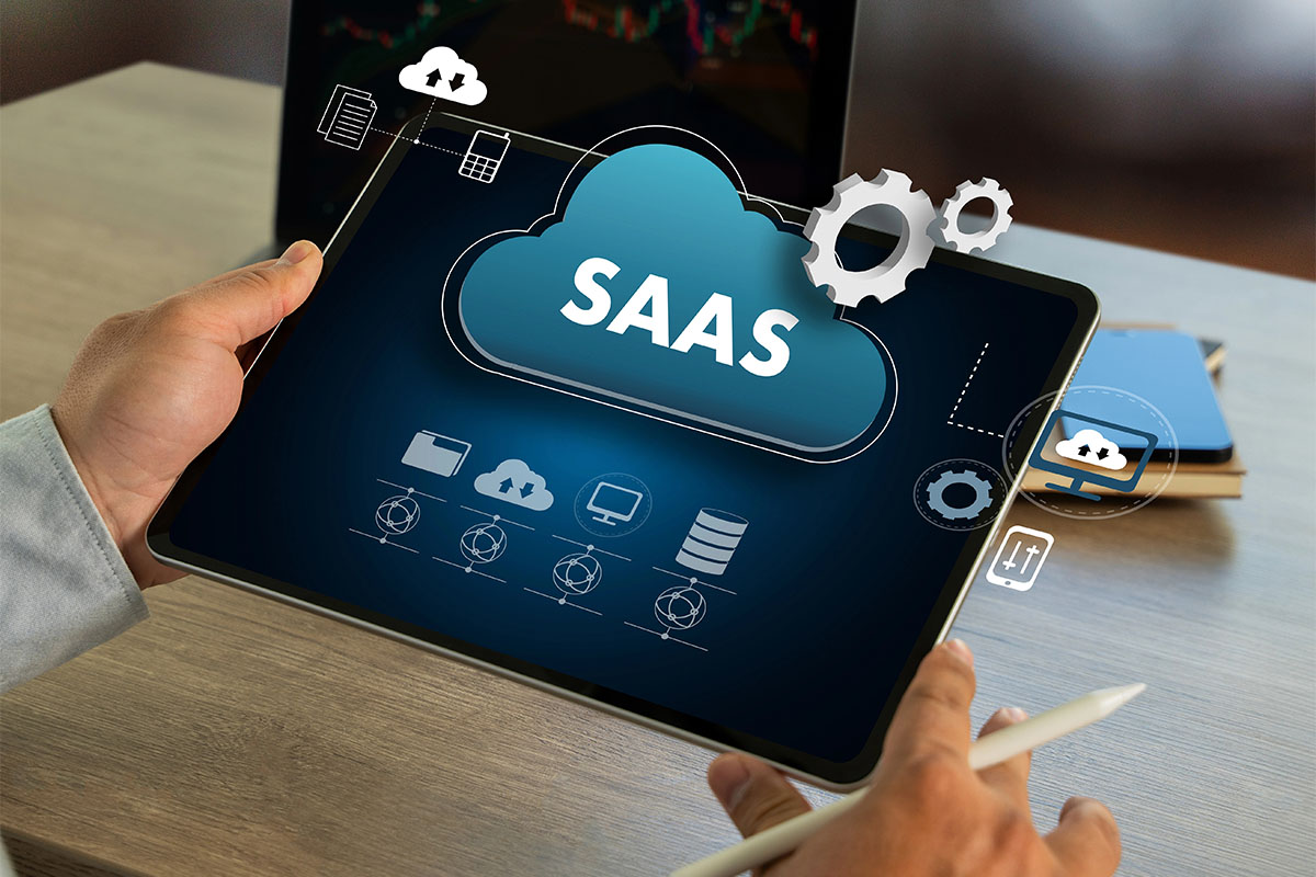 A person holding a tablet displaying a graphic representation of "SAAS" within a cloud, surrounded by icons symbolizing data transfer, storage, and software functionality on a wooden desk.