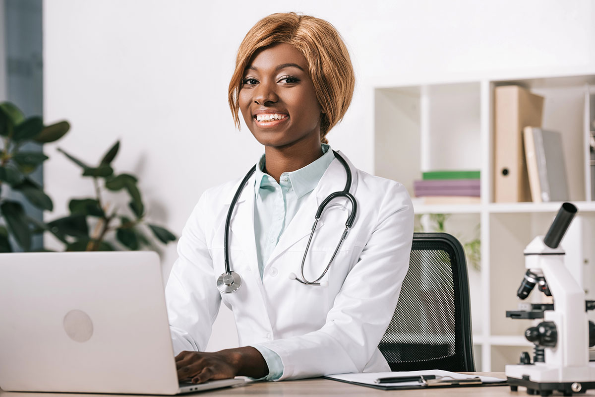 A smiling female doctor in a white lab coat with a stethoscope around her neck sits at a desk with a laptop, surrounded by indoor plants and a microscope in the foreground.