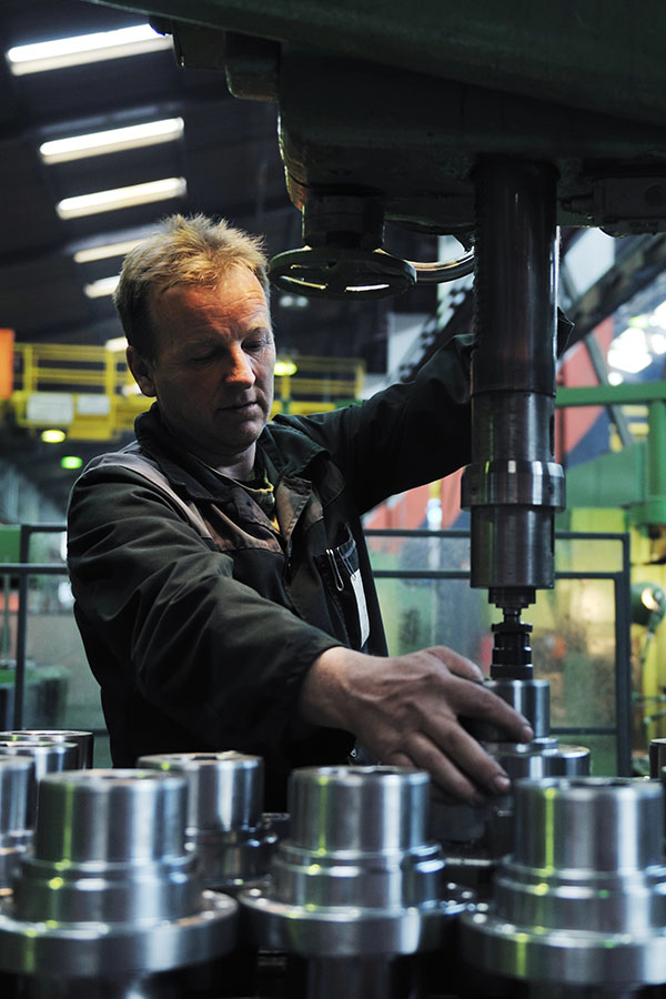 A worker in a factory operates machinery, focusing on assembling metal components. Bright industrial lighting illuminates the space, highlighting the organized arrangement of shiny metal parts in the foreground.