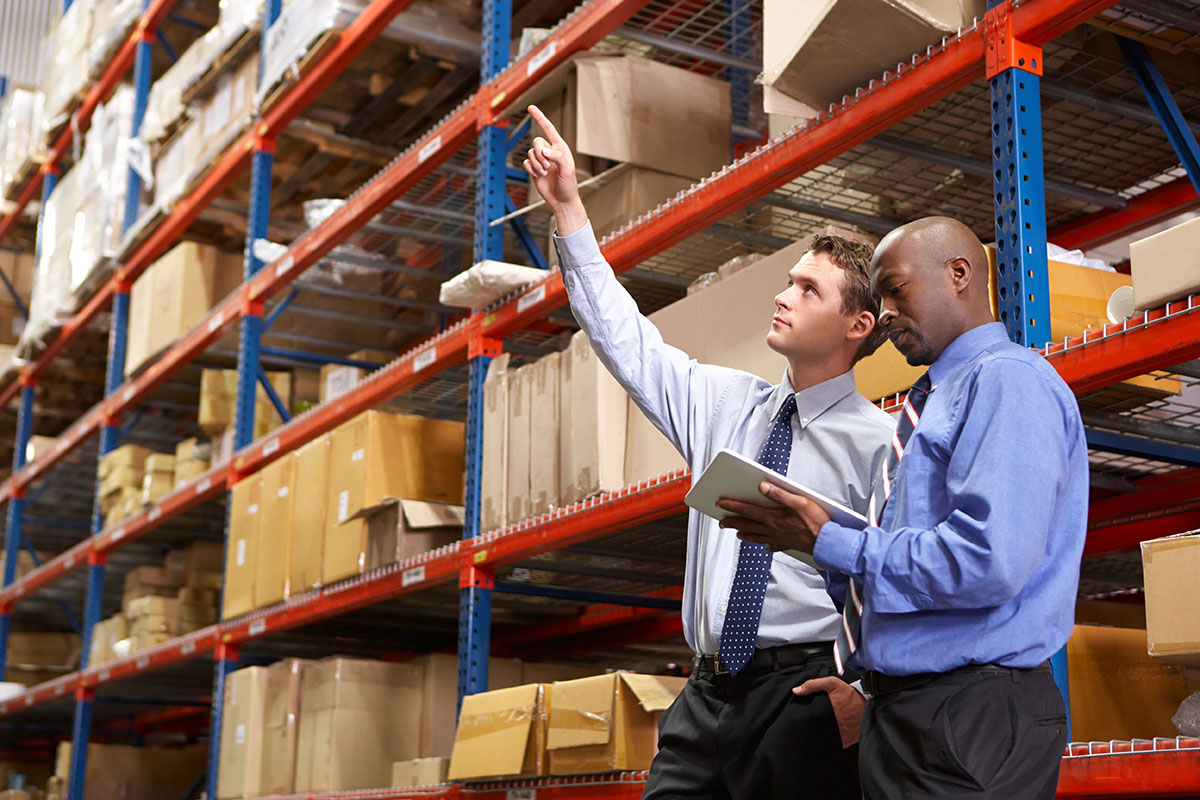 Two men in business attire are discussing items in a warehouse, with one pointing at a higher shelf while the other holds a tablet and looks on.